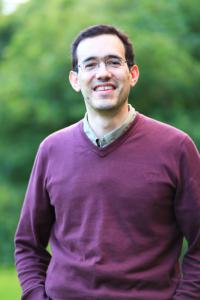 A man in a burgundy jumper smiles at the camera. He is against a blurred nature backdrop.