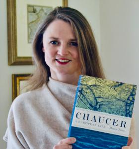 Smiling woman faces camera. She is holding a copy of her book 'Chaucer; A European Life', is against a white background, and is wearing a cream jumper