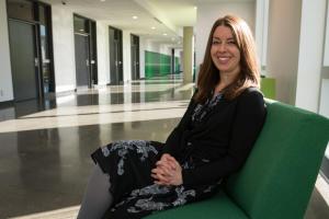 Smiling woman faces the camera, seated upon a green chair in a long corridor at an academic institution. She is dressed in a black blazer and patterned dress.