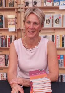 Woman smiles at the camera. She is in a pink top and wearing a necklace. In front of her is a pile of books, and in one hand is a pen.