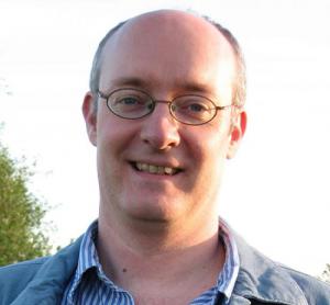 A man smiles at the camera. He is wearing thinly rimmed glasses, a light jackets, and a blue and white striped shirt.