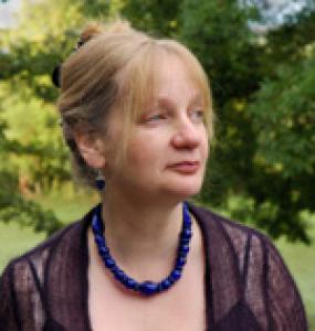 profile of blond-haired woman wearing a sheer black top and dark blue necklace. Trees in background