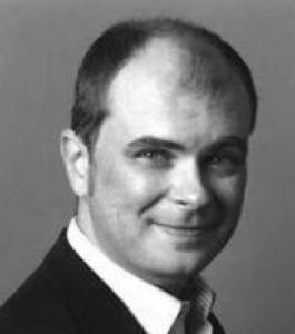 Close up black and white image of a man smiling with his mouth closed, facing the camera. He is in a black coat and white shirt.