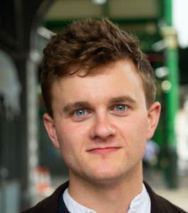 A man faces the camera. He has a black coat and white shirt on