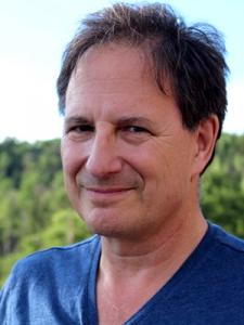 A man faces the viewer. He is in a bue t-shirt and is outside, in a natural backdrop.