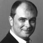 Close up black and white image of a man smiling with his mouth closed, facing the camera. He is in a black coat and white shirt.