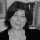 A black and white photo shows a headshot of a woman staring at the camera. She is wearing a black top and is stood in front of a bookshelf.