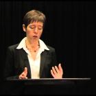 A woman in a black coat and white shirt is giving a lecture. Her hands are animated, and she is standing in front of a black curtain.