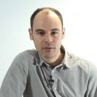 Man stares at the camera, against a light blue backdrop. He is wearing a grey shirt. 